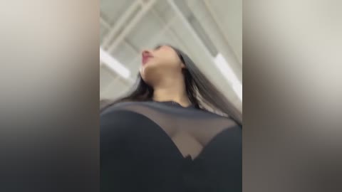 A video showing a close-up of a woman with long black hair, wearing a sheer black top with a plunging neckline, set against a modern industrial background with exposed beams and fluorescent lighting.