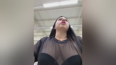 Media: Video of a woman with long dark hair, wearing a sheer black top, her breasts partially visible, standing in an industrial setting with high ceilings and pipes.