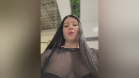 Media: Video of a young Latina woman with long black hair and medium skin tone, wearing a sheer black top, standing indoors with a blurred green plant and ceiling lights in the background.