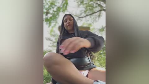 Media: A video of a woman in a black hoodie, sitting on a lawn, her hand touching a tree branch above her head. The background shows a blurred white building and green foliage.