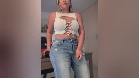 Media: Video of a blonde woman with fair skin, wearing a cropped white tank top with a lace-up front and high-waisted blue jeans, standing in a modern kitchen.