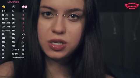 A close-up video of a young woman with long, dark hair, light skin, and a neutral expression, taken indoors with a dark background. The image includes a social media interface on the left.