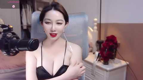 Media: Video of an attractive East Asian woman with light skin and dark hair, wearing a black bra, recording herself with a camera in a modern bedroom with a white nightstand and red roses.