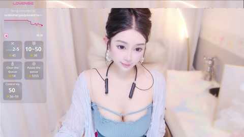 Media: Video of a young East Asian woman with fair skin, dark hair, wearing a light blue top and headphones, sitting in a softly lit bedroom with white curtains and a bed.