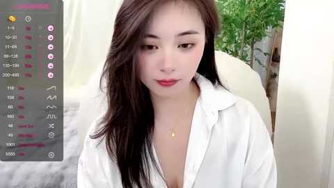 Media: Video of an East Asian woman with long black hair, fair skin, and red lipstick, wearing a white blouse, indoors with green plants and a white bed.