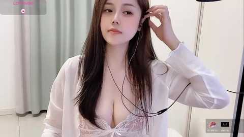 Media: Video of an Asian woman with long, straight black hair, wearing a white robe, revealing a lace bra, adjusting headphones in a modern, minimalist room with white walls and light-colored curtains.