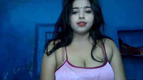 Media: Video of a young woman with medium skin tone, wearing a pink camisole, standing against a blue wall. She has long, dark hair, red lipstick, and a bindi on her forehead. Background shows a blurred room with shelves.