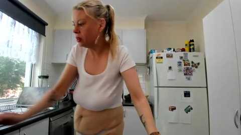 Media: Video of a blonde, fair-skinned woman with large breasts and a tattoo on her arm, wearing a white crop top and beige pants, cooking in a modern kitchen with white cabinets and a cluttered fridge.