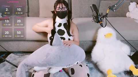 Media: Video of an Asian woman with fair skin, long black hair, wearing a cow-patterned onesie, black mask, and glasses, sitting on a gray couch with a stuffed duck.