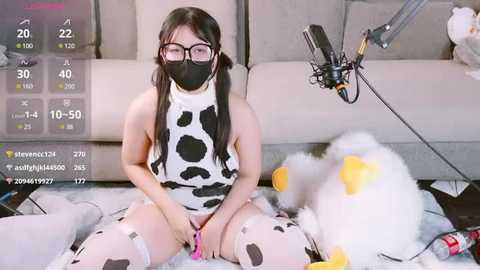 Media: Video of an Asian woman in cow-themed lingerie, sitting on the floor with a stuffed duck, wearing a face mask, surrounded by plush toys.