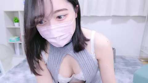 Media: Video of a young Asian woman with straight black hair and light skin, wearing a face mask and a grey ribbed top, revealing a white lace bra. She's in a bright, minimalist room with white walls and light blue furniture.