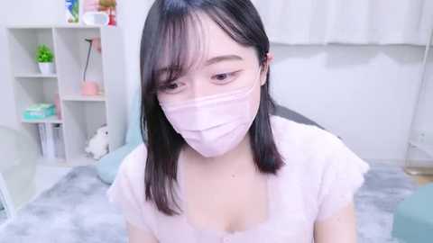 Media: Video of an Asian woman with straight black hair and light skin, wearing a white face mask, light pink top, and a grey blanket. Background shows a white room with a bookshelf and potted plants.