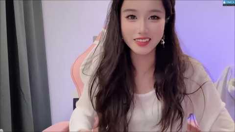 Media: Video of an East Asian woman with long, wavy black hair, fair skin, and delicate features, wearing a white top, smiling, sitting in a gaming chair, with a purple wall and gray curtains in the background.