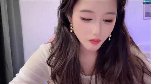 Media: Video of a young Asian woman with long, wavy black hair, fair skin, and full lips, wearing a white top and dangling earrings, looking down pensively in a softly lit room.