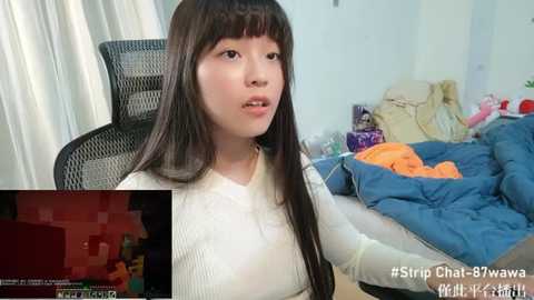 Media: Video of an Asian woman with long black hair, wearing a white sweater, sitting on a black chair in a messy bedroom with a blue quilt and colorful clothes.