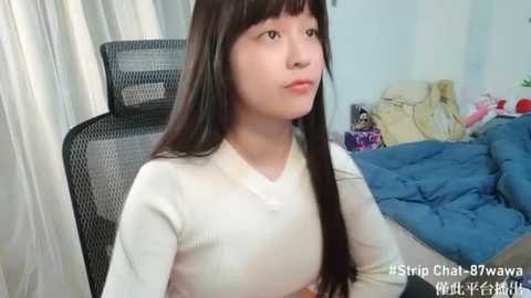 Media: Video of a young Asian woman with long, straight black hair and fair skin, wearing a white long-sleeve top, sitting on a black office chair in a cluttered bedroom with a bed and piles of clothes.