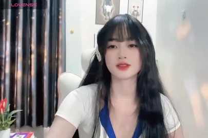 Media: A video of an East Asian woman with long black hair and fair skin, wearing a white blouse with blue trim, sitting in a brightly lit room with dark curtains and a potted plant.