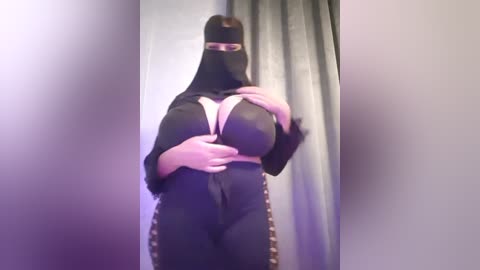 Media: A video of a plus-sized woman in a black nun costume with a white cross, standing against a grey curtain. Her ample breasts are accentuated by the tight outfit.