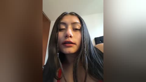 Media: A video of a young woman with long, dark hair, light skin, and full lips. She's indoors with a beige wall and wooden door in the background, looking slightly off-center.