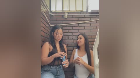 Video of two young women with light skin, one in a black tank top and jeans, the other in a white tank top, sitting on brick steps, holding a bottle.