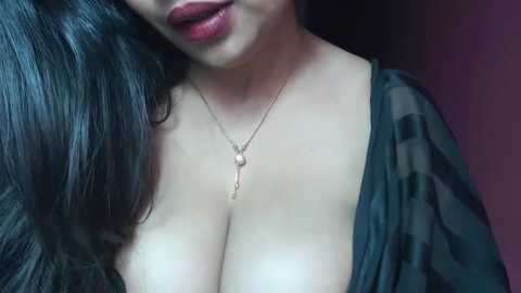 Media: Video of a woman with long, dark hair and fair skin, wearing a low-cut, black sheer dress revealing ample cleavage. She has full, glossy red lips and a delicate gold necklace with a pendant.