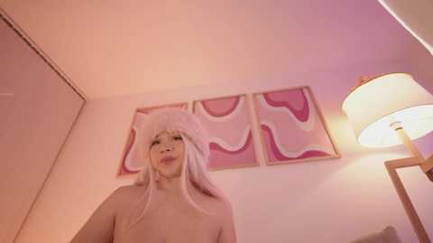 Media: Video of an Asian woman with light skin, long straight hair, wearing a cat headband, topless, standing in a cozy, pink-lit room with a modern abstract painting and a lamp.