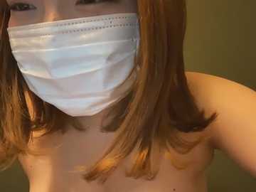 Media: A close-up video of a woman with shoulder-length, auburn hair, wearing a white surgical mask, revealing her bare shoulders. The background is blurred, featuring muted green and beige tones.