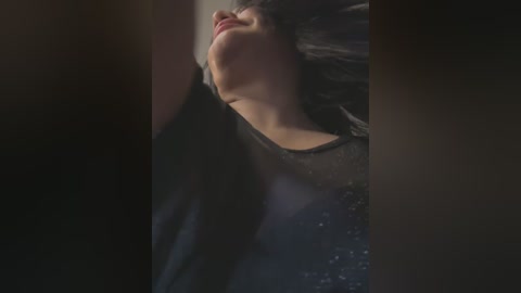 Media: A close-up video of a person with fair skin, long black hair, and closed eyes, lying on a dark, textured surface. The image is dimly lit, emphasizing a serene, introspective mood.