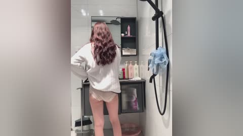 Media: Video of a woman with long brown hair, wearing a white robe and lacy white panties, standing in a modern, tiled bathroom with a glass shower, soap bottles, and a blue towel hanging on the wall.