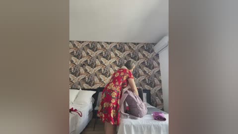 Media: Video of a woman in a red floral dress bending over a bed, adjusting a pink bag. The room features a patterned wallpaper and a white bed with pillows.