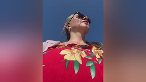 Media: Video of a blonde woman wearing sunglasses, a red dress with large yellow flower design, and a pink shoulder bag, gazing upward at a clear blue sky.