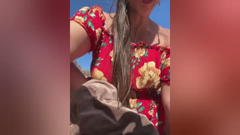 Media: Video of a woman with wet, dark hair, wearing a red floral top, holding a black and gray towel, against a bright blue sky background.