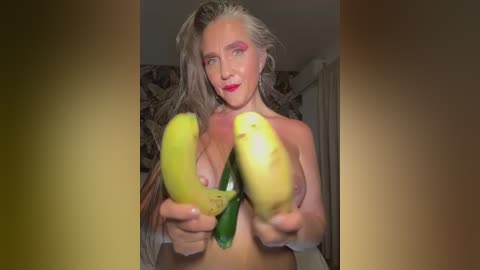 Media: Video of a topless, fair-skinned woman with long, silver hair, holding two bananas in front of her chest, against a patterned wall background.