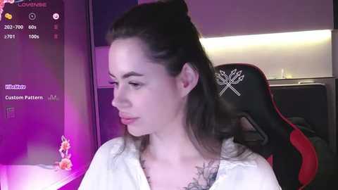 A video of a fair-skinned woman with long brown hair, wearing a white robe, sitting in a red and black gaming chair, in a dimly lit room.