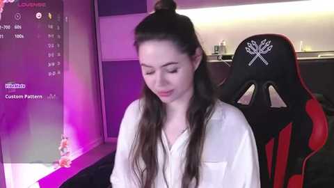 Media: Video of a young woman with long dark hair tied up in a bun, wearing a white shirt, sitting in a black and red gaming chair in a dimly lit room.