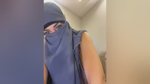 Media: Video of a woman with medium brown skin, wearing a blue hijab, peering out from behind a black curtain, revealing dark eyes. She has a sleeveless top and beige walls in the background.