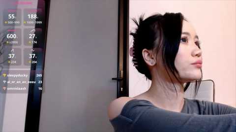 Video of a young woman with fair skin and black hair styled in a messy bun, wearing a grey off-shoulder top, standing in a room with a calendar screen displaying dates and statistics.