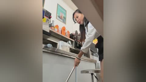 Media: A video of an Asian woman in a white chef's uniform cleaning a stainless steel kitchen counter with a cloth. The background includes a wall-mounted clock, framed art, and orange utensils.