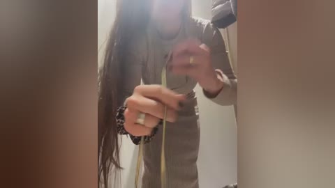 Media: Video of a woman with long, straight brown hair, wearing a grey long-sleeve shirt, measuring her waist with a yellow measuring tape in a dimly lit room.