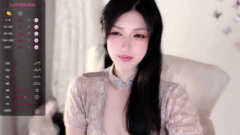 Media: Video of an East Asian woman with long black hair, wearing a beige lace top, sitting on a white chair. The background is blurred, showing a room with white walls and a hint of a bed.