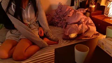 Video of a woman in a grey hoodie and shorts, sitting on a striped bed with a pink blanket, surrounded by cups and a warm, cozy room.