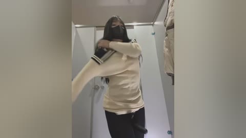 Media: Video of a woman in a beige sweater and black pants, wearing a mask, swinging a black jacket in a narrow hallway with white walls and a closed door.