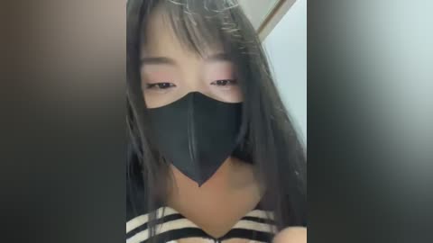 Media: A close-up video of an Asian woman with long black hair, wearing a black face mask and a striped top. She has light skin and is indoors, likely in a bathroom.