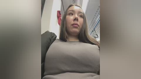 Media: A video of a plus-sized woman with long brown hair, wearing a gray sweater, sitting in a subway car. The background features a tiled wall and a red emergency sign.