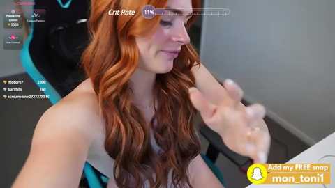 Video of a fair-skinned woman with long, wavy red hair, wearing a strapless top, sitting in a gaming chair. She gestures with her hand while looking at a computer screen displaying Twitch chat.