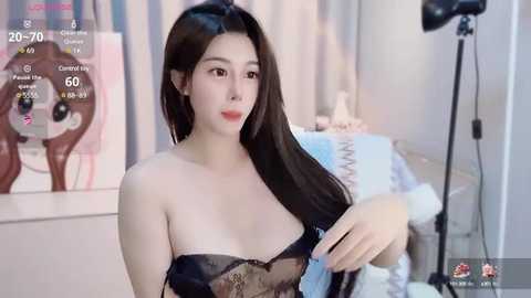 Media: Video of an East Asian woman with long black hair, fair skin, wearing a black lace bra, standing in a softly lit bedroom with a large teddy bear and a camera on a tripod.