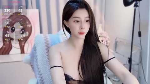 Media: Video of an East Asian woman with long black hair, wearing a black lace bra, sitting in a brightly lit bedroom with striped curtains and a white bed.