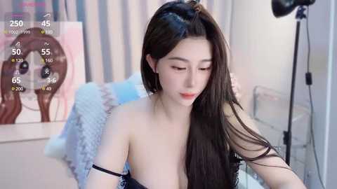 Media: Video of a young Asian woman with long black hair, fair skin, and light makeup, wearing a black lace bra, sitting in a modern room with a striped curtain and a pink abstract painting.