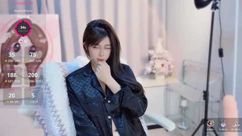 Media: Video of an East Asian woman with long black hair, wearing a black patterned jacket, sitting on a bed with a floral duvet. Background features a light-colored room with a vase of flowers and a lamp.