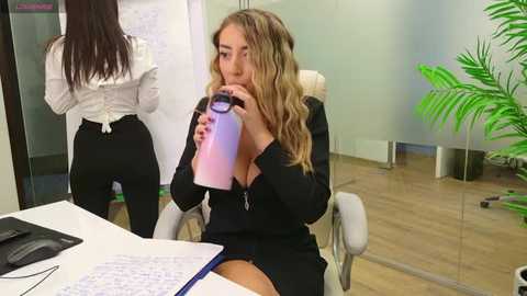 Media: Video of a blonde woman in a black suit drinking from a pink water bottle, sitting at a desk in an office with glass walls and a green plant.